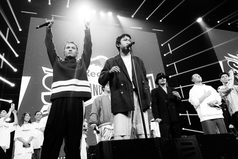 A black and white shot captures a group of performers on stage, bathed in bright spotlights, with a large screen displaying the 'Red Bull Sound Select' logo in the background.