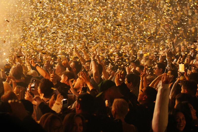 A crowd of people is cheering with their hands up as confetti falls from above.