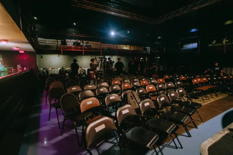 The image shows rows of empty chairs in a dimly lit event space, with camera operators setting up in the background.