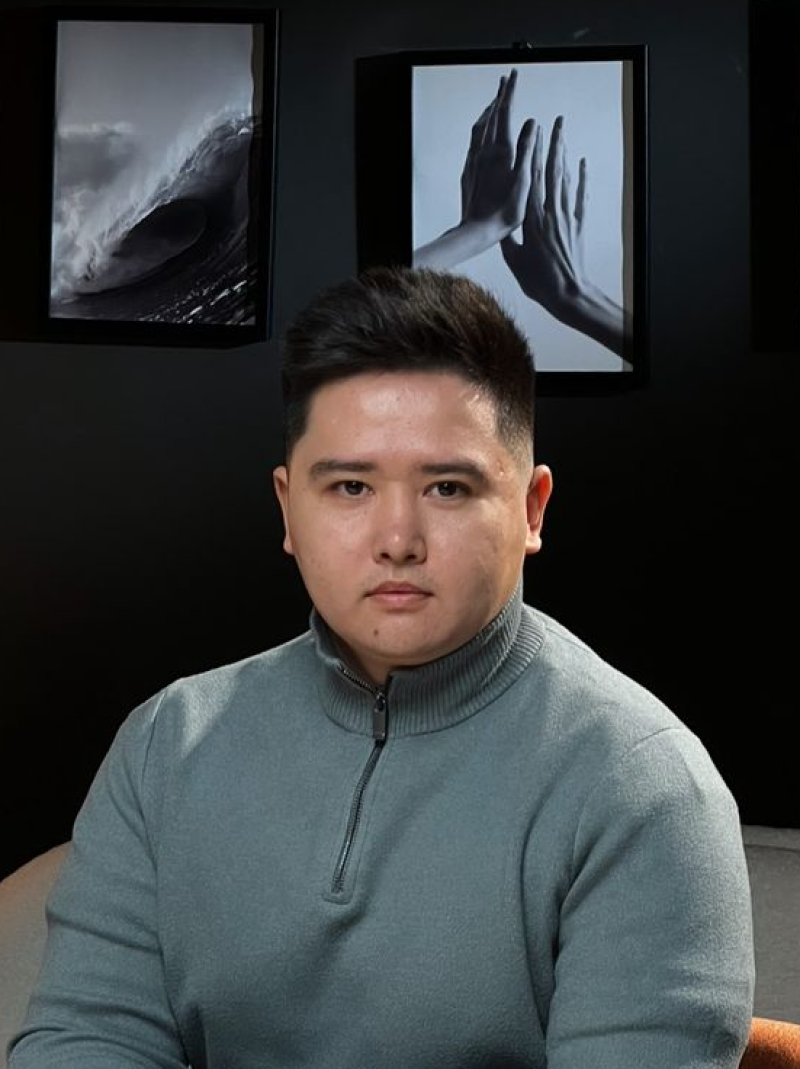 A man wearing a gray turtleneck sweater with a zipper poses in front of a black wall with two framed pictures: one of a wave and the other of hands touching.