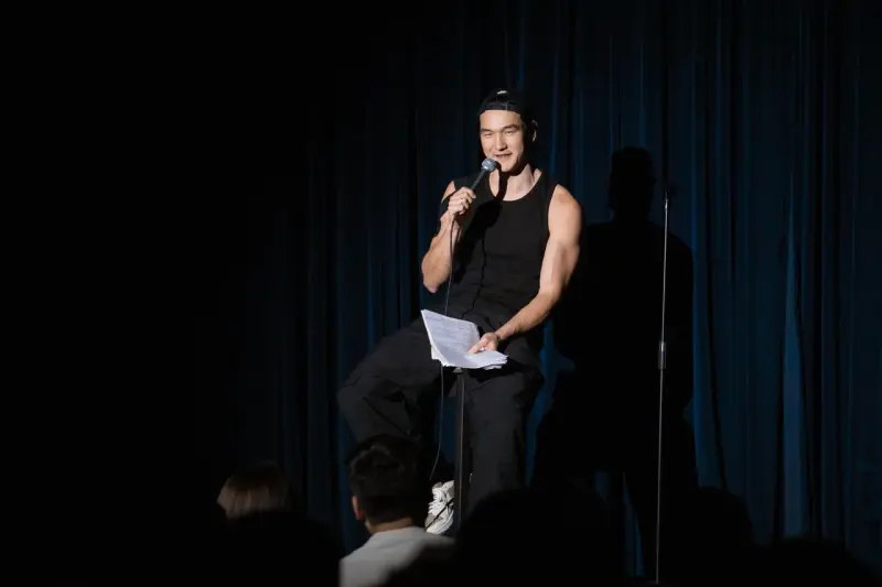 A man in a black tank top and cap sits on a stool on stage, holding a microphone and papers, in front of a blue curtain, with audience members in the foreground.