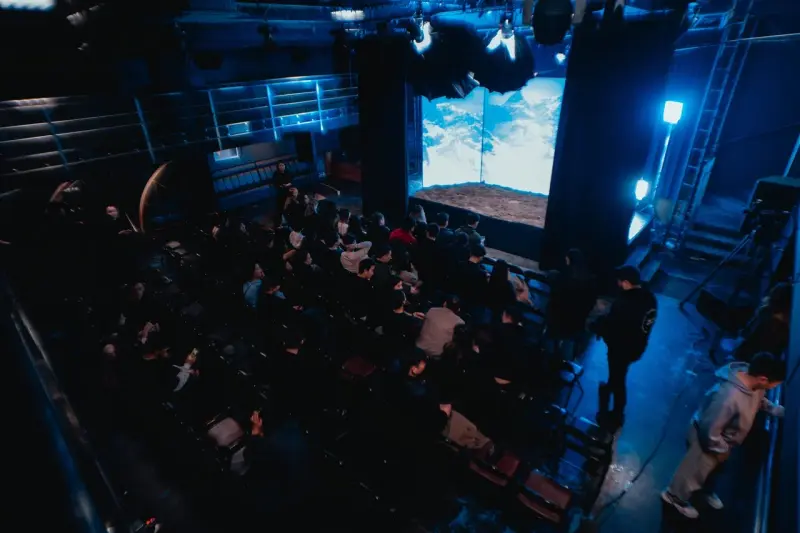 A theater filled with an audience watching a screen displaying a sky and ground scene, lit with blue lighting.