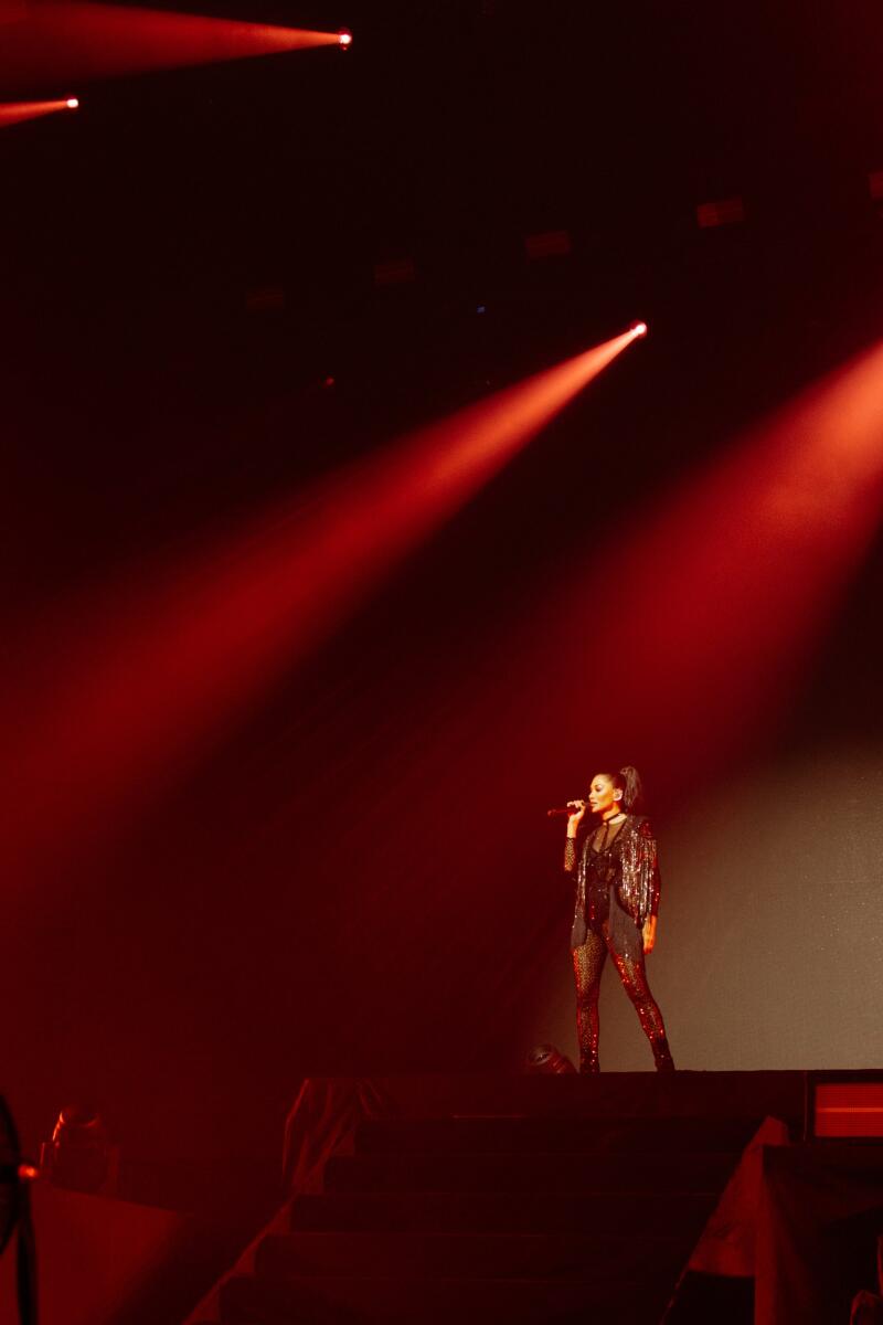 A singer in a sequined outfit performs on stage with a microphone under red stage lights.