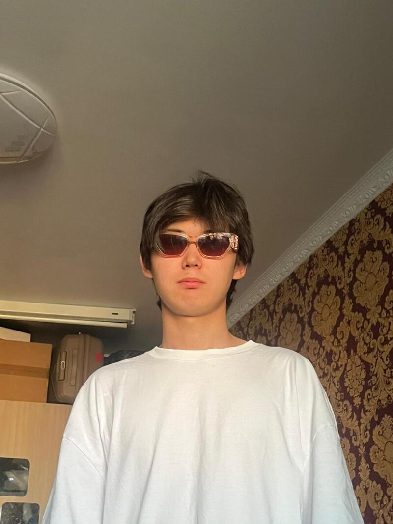 A young person with dark hair and light-framed sunglasses, wearing a white t-shirt, looks directly at the camera. The background features patterned wallpaper, a ceiling light, and furniture.