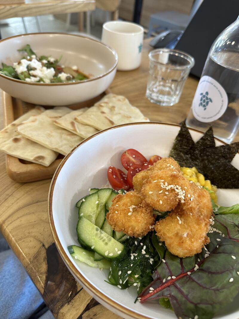 The image shows a wooden table with two bowls of food, flatbread, a glass, and a water bottle. One bowl contains a salad with fried food, cucumbers, tomatoes, corn, and seaweed; the other bowl has greens and white cheese. There are also flatbreads on a wooden board.