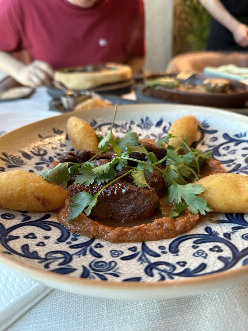 The image shows a plate of food with meat, sauce, fried potatoes, and cilantro, with people and other food in the background.