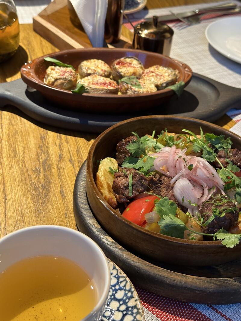 The image shows two dishes and a cup of tea on a wooden table: the first dish contains small, round foods covered with a sauce, while the second is a wooden bowl filled with meat, potatoes, and vegetables, garnished with cilantro and red onions. The tea is in a white cup with a blue pattern.