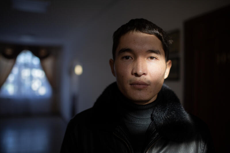 Крупный план мужчины в черной куртке с меховым воротником, половина лица которого освещена солнечным светом.