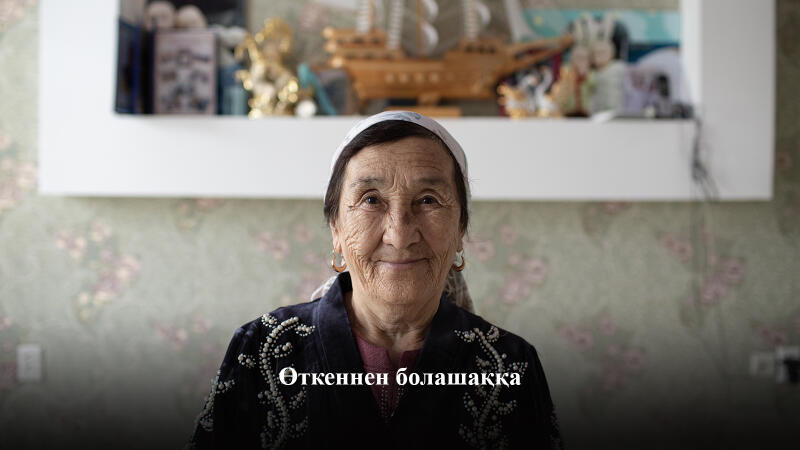 Пожилая женщина в платке и серьгах слегка улыбается в камеру, с казахским текстом "Өткеннен болашаққа" внизу изображения.