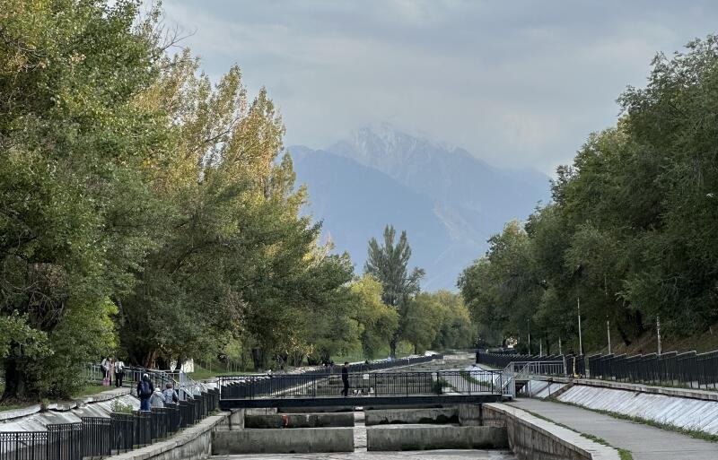 The landscape features a concrete canal with a series of dams, framed by a footpath with a black metal fence, and lush trees on either side, with mountains visible in the background under a cloudy sky.