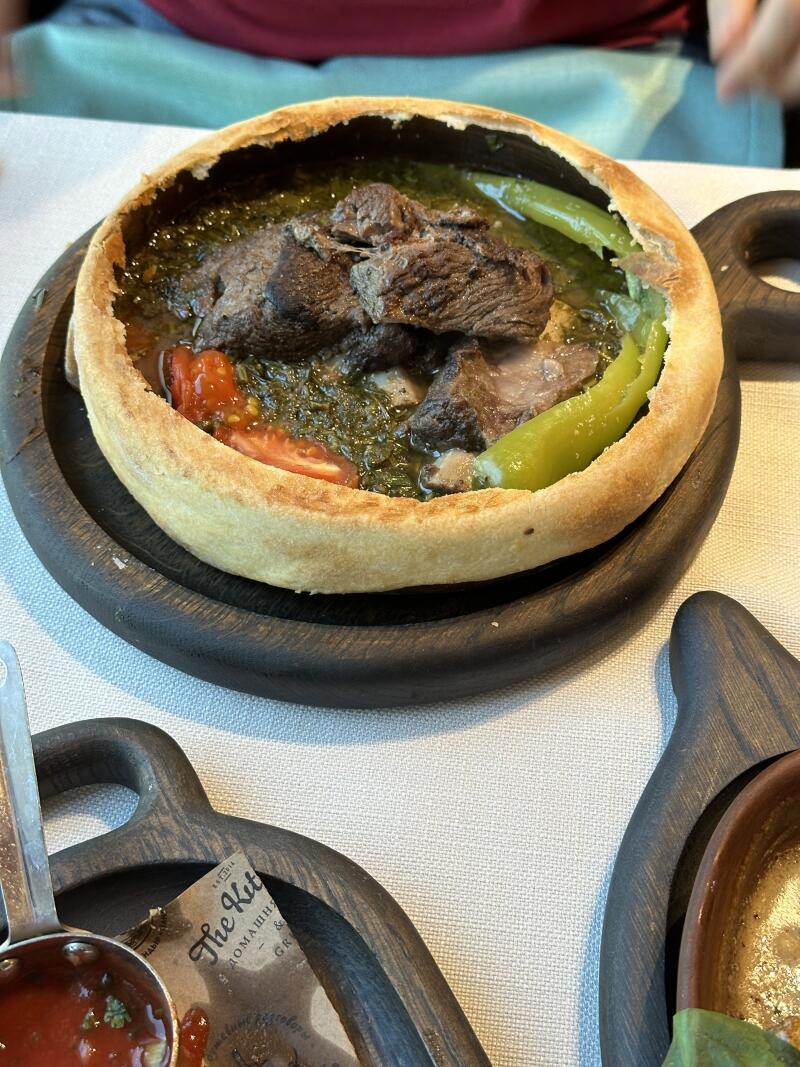 A dish featuring meat, green peppers, and tomato, served in a bread bowl on a dark wooden plate.