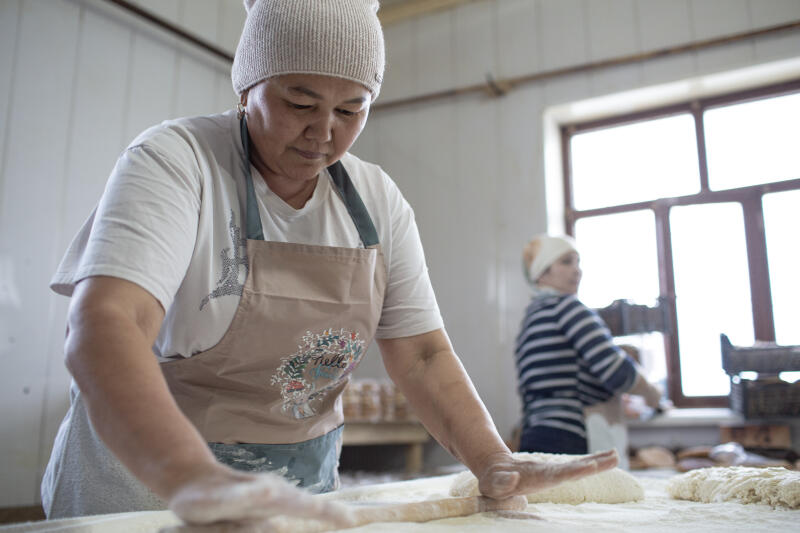 В пекарне женщина в белой шапке и фартуке раскатывает тесто скалкой на посыпанной мукой поверхности, на заднем плане видна другая женщина.