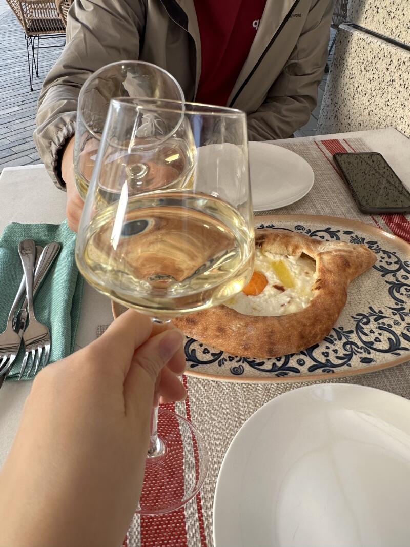 A table setting featuring two glasses of white wine, a traditional Adjarian khachapuri, plates, cutlery, and a phone.