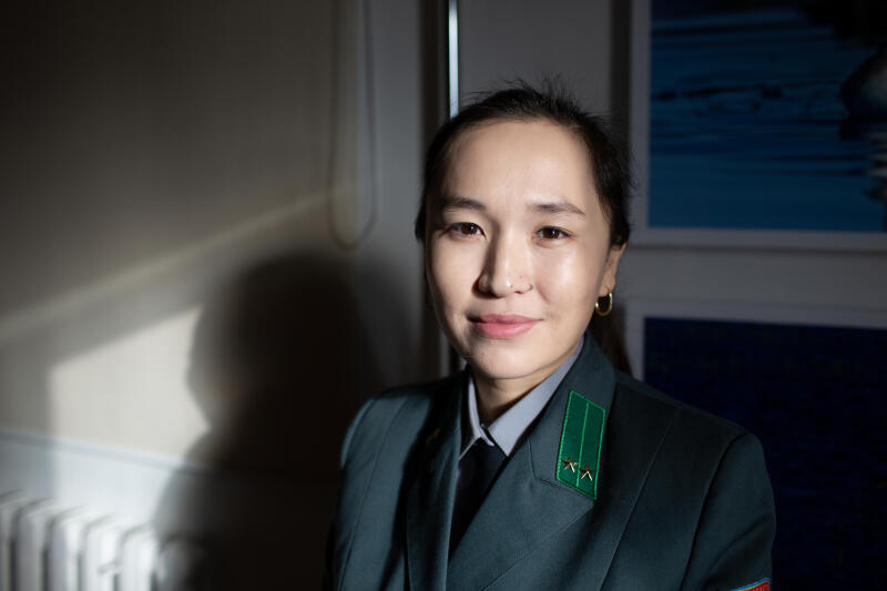 Женщина в темно-зеленой военной форме с двумя звездами на погонах стоит на фоне белой стены.