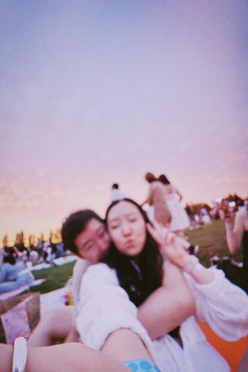 A couple poses for a selfie at an outdoor event; the woman is making a peace sign with her hand, and there is a crowd of people in the background against a pink and blue sky.