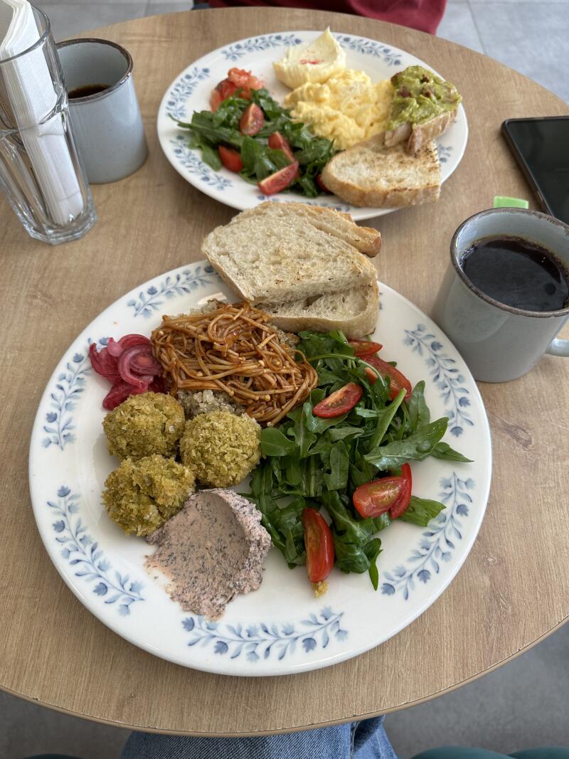 The photo shows two plates of food on a wooden table. One plate contains a salad with tomatoes and arugula, falafel, pickled onions, some noodles, hummus, and bread. The other plate contains scrambled eggs, hummus, and a salad with tomatoes and arugula, and bread. There are also two mugs of coffee and a glass with napkins on the table.