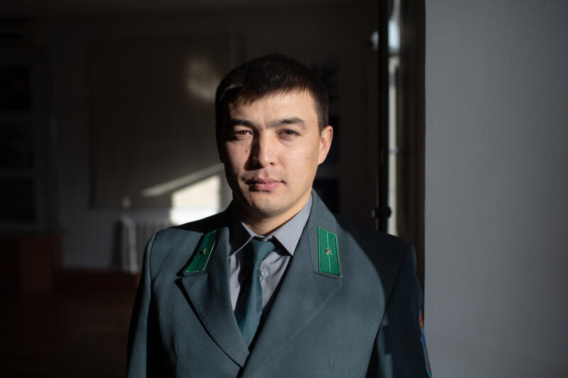 Мужчина в тёмно-бирюзовом костюме с зелёными погонами стоит в комнате с солнечным светом.