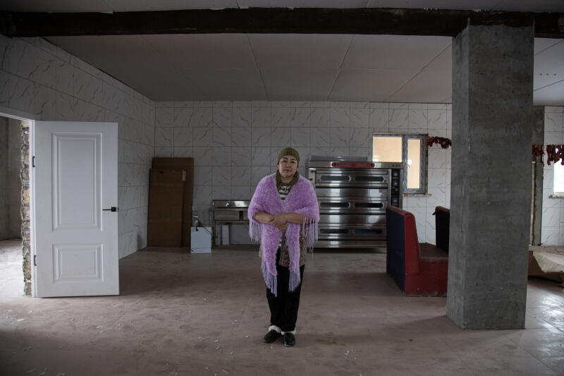 Женщина в лавандовой шали стоит в центре большой, недостроенной комнаты с облицованными плиткой стенами, большой духовкой и белой дверью.