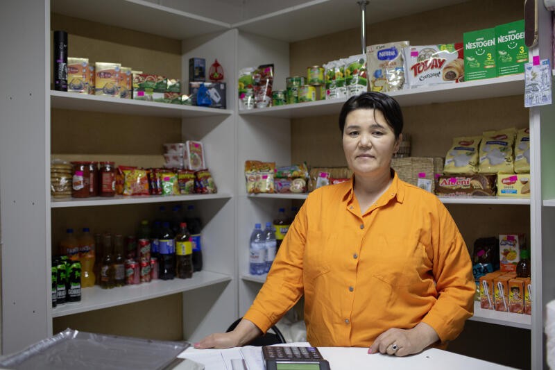 Женщина в оранжевой рубашке стоит за прилавком в небольшом магазине, заполненном полками с различными продуктами.