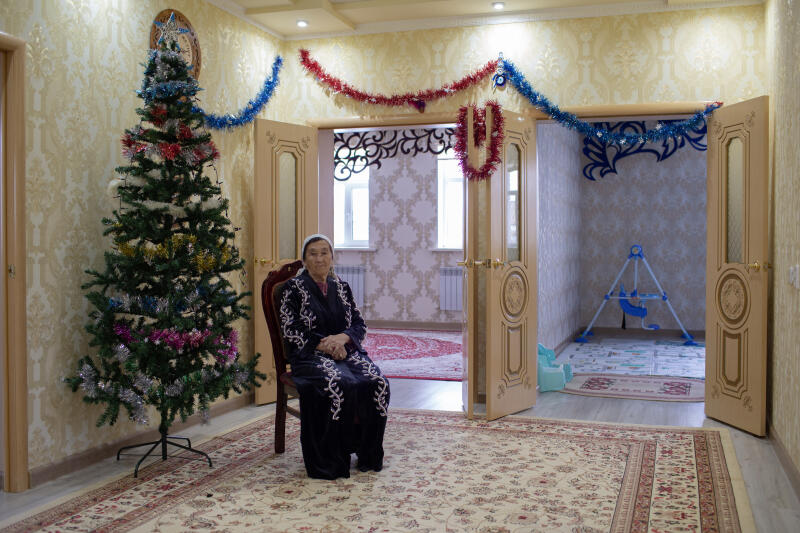 Пожилая женщина сидит на стуле рядом с новогодней елкой в комнате, украшенной гирляндами.