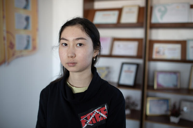 Портрет молодой азиатской женщины с темными волосами, одетой в черную рубашку с нашивкой, на которой написано "THE TEMPEST FIRE", стоит перед полкой с рамками, заполненными сертификатами.