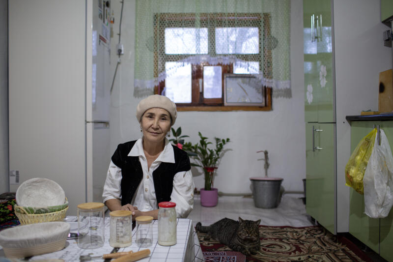 Женщина в бежевом берете сидит за столом с принадлежностями для выпечки хлеба, а полосатый кот лежит на ковре поблизости.