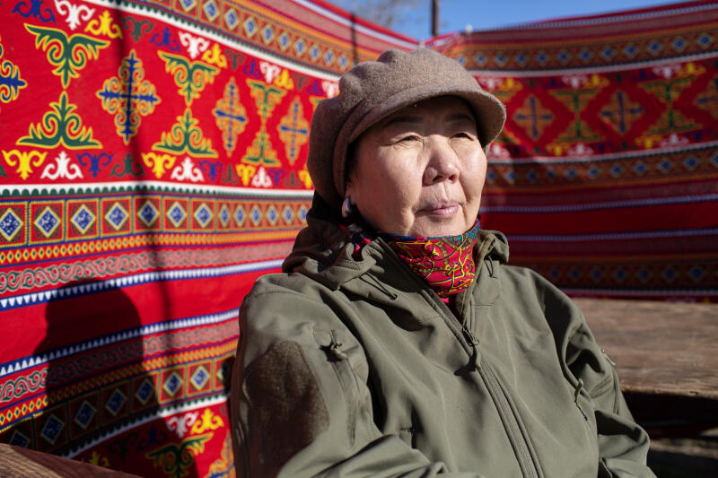 Женщина в коричневой кепке и зеленой куртке сидит на фоне красной стены с узорами.