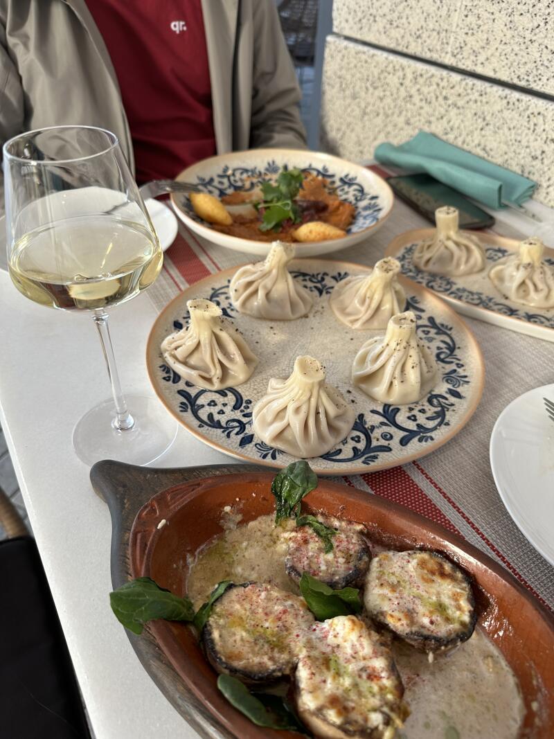 The image shows a table setting with various dishes: a glass of white wine, plates of khinkali (Georgian dumplings) and stuffed mushrooms, and another dish with sauce and dumplings, all arranged on a striped tablecloth.