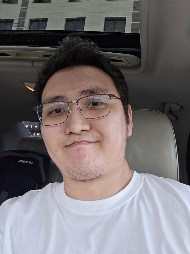 A selfie of a man with glasses and a white T-shirt in a car, with a building visible through the sunroof.