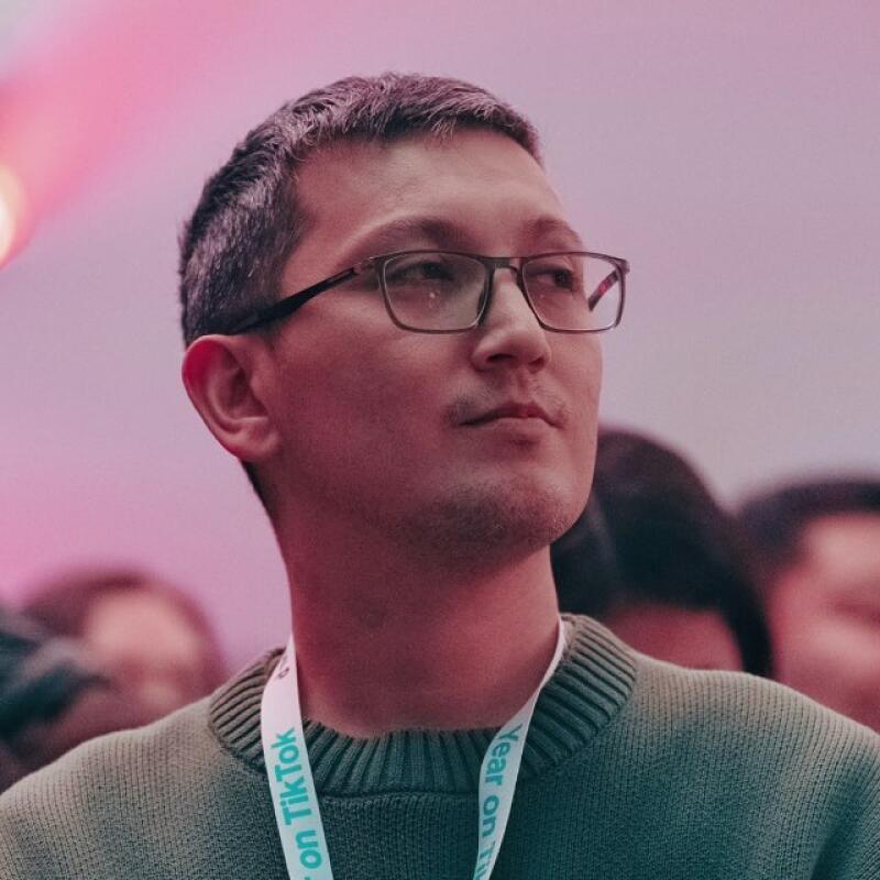 A man with short grey hair and glasses, wearing a green sweater and a white lanyard with "on TikTok" text, looks up and to the right against a blurred pink background.