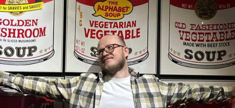 A man with glasses and a beard stands in front of three large soup can labels that read, "GOLDEN MUSHROOM SOUP", "THE ALPHABET SOUP VEGETARIAN VEGETABLE", and "OLD FASHIONED VEGETABLE SOUP."