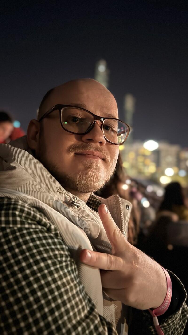 A man with glasses and a beard is making a peace sign and wearing a pink wristband that says "SEATING". He is wearing a gray jacket and a checkered shirt.