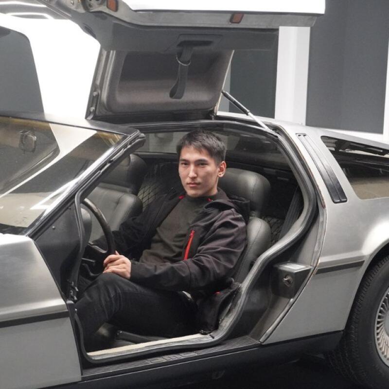 Мужчина сидит на водительском сиденье автомобиля DeLorean с открытой дверью типа "крыло чайки".