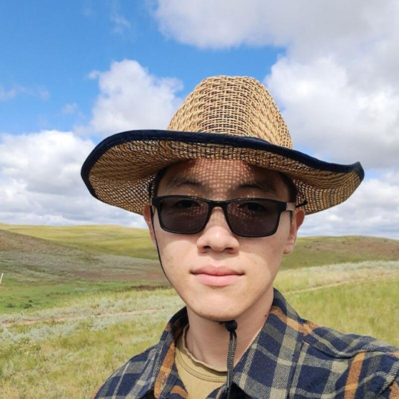 A person with sunglasses and a straw hat is standing in a field, wearing a plaid shirt.