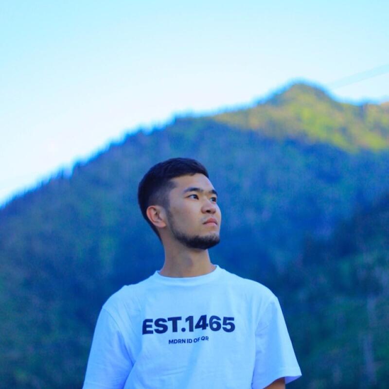 Молодой человек с бородой в белой футболке с надписью "EST.1465 MDRN ID OF QR" стоит на улице на фоне гор и светло-голубого неба.