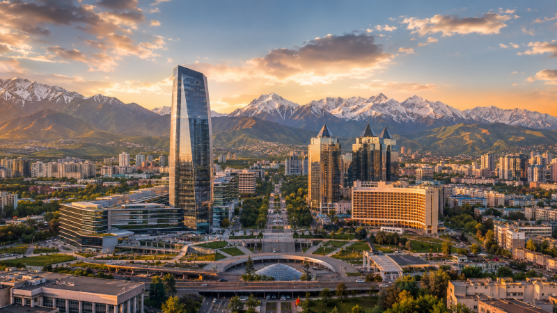 An aerial view of a modern city featuring skyscrapers and green boulevards, with majestic snow-capped mountains in the background under a colorful sunset sky.