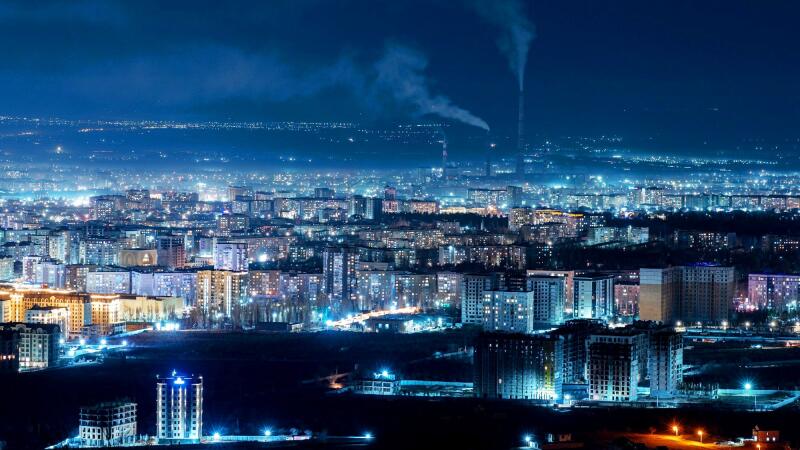 An aerial view shows a cityscape at night with illuminated buildings and smoke rising from distant chimneys.