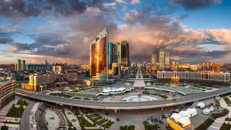 This is a panoramic view of a city with modern architecture, featuring tall buildings, a central plaza with a fountain, and a complex road system under a sky with dramatic clouds.