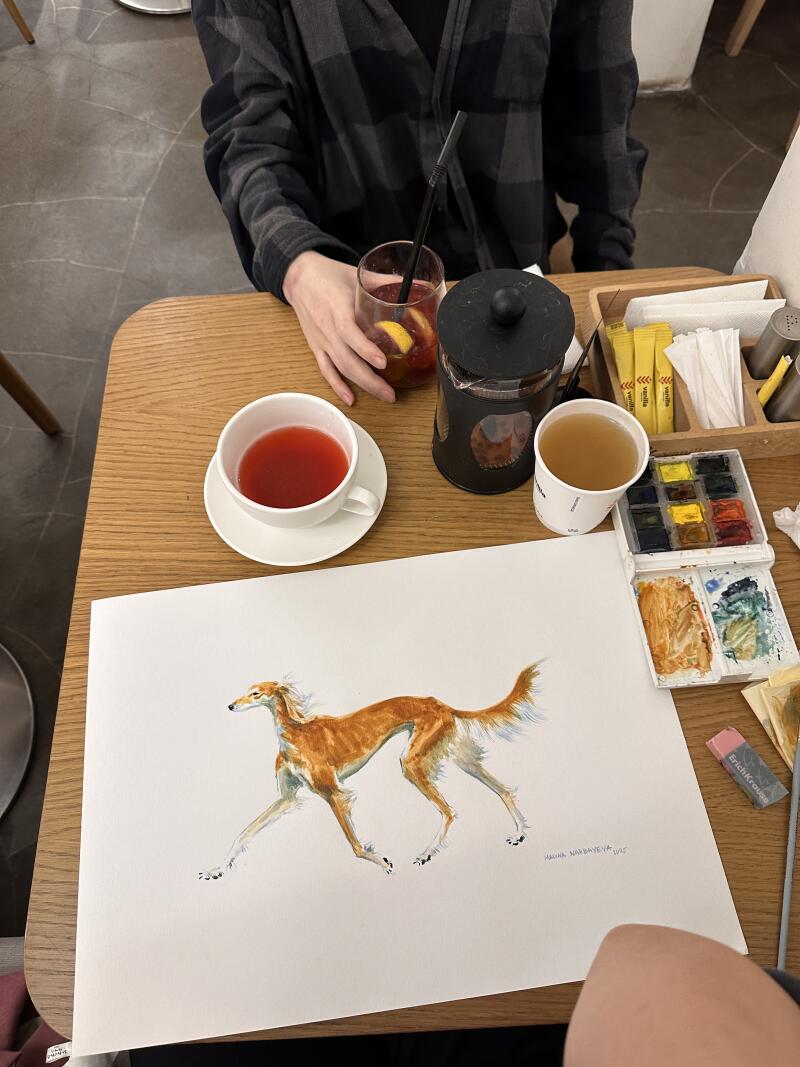На деревянном столе стоит рисунок собаки, рядом чашки с чаем, напиток с соломинкой и набор акварельных красок.